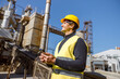 Joyful male worker in safety helmet holding cellphone and smiling while standing outdoors at factory