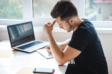 Side view of a businessman writing something in his notebook using mobile smart phone while working on laptop computer with digital tablet on desk.