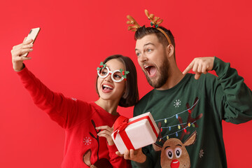 Happy young couple in stylish Christmas clothes and with gift taking selfie on color background