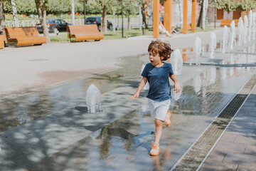 little boy is running in the street and playing with the water jets of a fountain spouting from the ground. child in blue T-shirt and white shorts is fooling around outdoors. lifestyle. space for text