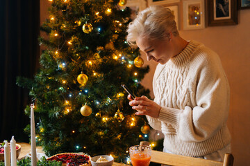 Wall Mural - Side view of pretty blonde young woman taking photo or making video of beautiful Christmas dinner table on smartphone, on blurred background of decorating xmas tree and celebration bokeh lights.