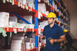 © chokniti - Handsome young male employee concentrating and focusing on work wearing safety helmet and uniform in warehouse while standing between racks in aisle
