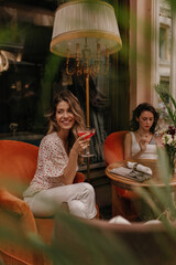 Wall Mural - Cute glamorous caucasian young ladies spend time sitting on red armchairs in restaurant with glass of wine. Brunette uses mobile phone away from her friend.