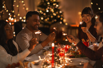 Wall Mural - holidays and celebration concept - multiethnic group of happy friends with sparklers having christmas dinner at home