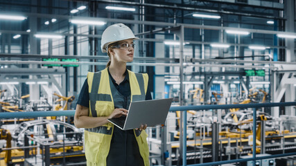 Fototapete - Portrait of Female Automotive Industry 4.0 Engineer in Safety Uniform Using Laptop at Car Factory Facility. Confident Assembly Plant Specialist Working on Manufacturing Modern Electric Vehicles.