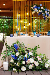 Wall Mural - Wedding presidium, banquet table for newlyweds with flowers, greenery, blue and gold color. Lush flower arrangement on the wedding table. Glowing light bulbs. Soft selective focus.