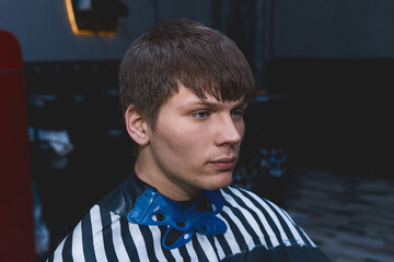 A shaggy untrimmed guy of Caucasian appearance with dark long hair sits in a chair in a hairdresser's client for a haircut. The need for hairdressing services, hair care