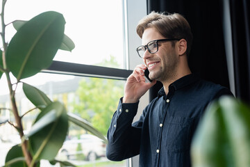 Positive programmer in eyeglasses talking on smartphone near plants in office