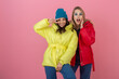 © mary_markevich - two attractive girl friends active women posing on pink background in colorful winter down jacket of bright red and yellow color having fun together, warm coat fashion trend, sincerely laughing