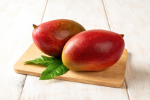 Two Red Ripe Palmer Mangoes On A Cutting Board Over White Wooden Table. Ready To Eat Whole Sweet Tasty Mango. Two Delicious Tropical Fruits Of Mangifera Indica Close-up.