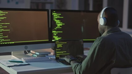 Rear-view stab shot of African-American programmer in headphones sitting in front of two monitors and laptop writing program codes, working late in empty office