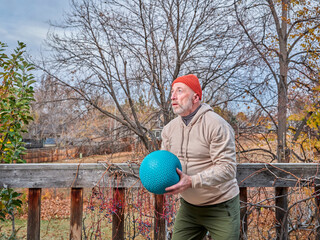 senior male (on late 60s) is exercising with a heavy slam ball in his backyard, chilly afternoon in fall scenery