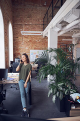 Full length shot of cheerful woman, architect or interior designer smiling away while talking on the phone, standing in modern green office