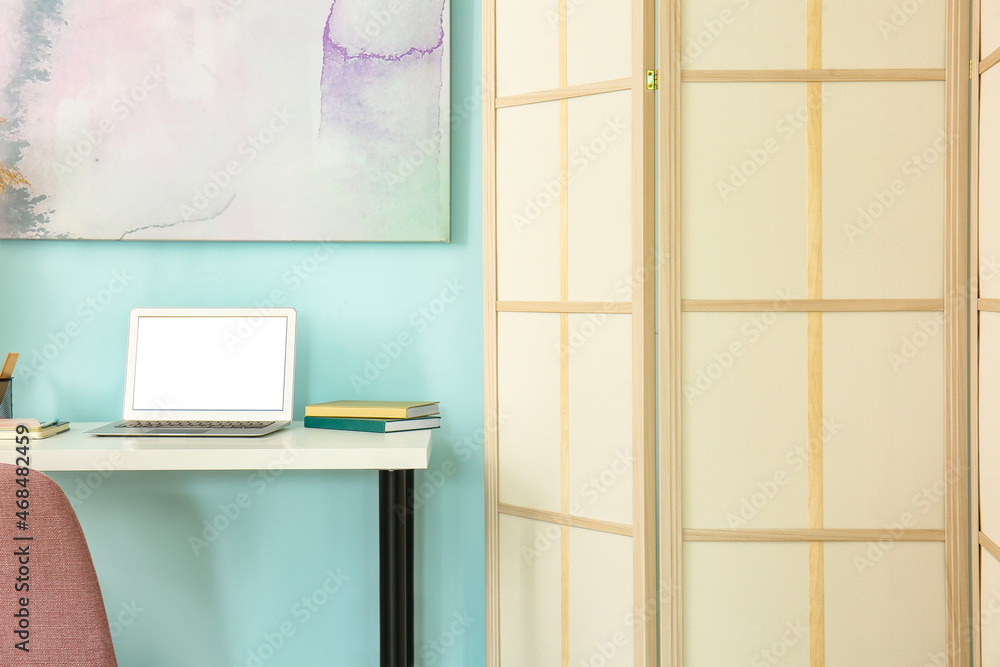 Modern workplace with laptop and folding screen near blue wall