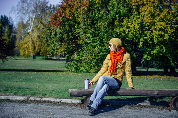 Wall Mural - Pretty european woman in yellow jacket and red scarf sitting on wooden bench in deserted park. Enjoying solitude and sunny autumn weather. Blurred plant background, copy space.