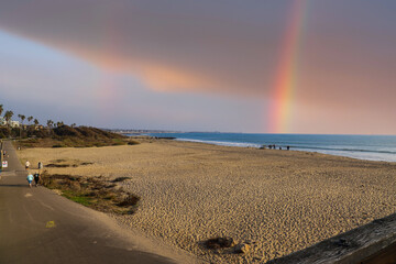 a rainbow over the beach with vast silky brown sand with people walking down the bike trail and people standing in the sand at Surfers Point at Seaside Park in Ventura California USA