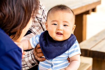 おばあちゃんに抱っこされてお母さんと戯れる赤ちゃんがカメラ目線　3世代が外のベンチに座っています