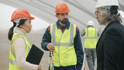 Wall Mural - Builders wearing uniform are talking at construction site while architect in suit and helmet is coming discussing project gesturing and looking around