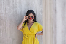 View Near A Column Of A Young Latina Woman In Yellow Dress Wearing Sunglasses And Holding Them With One Hand And Looking At Camera Over The Top Of The Glasses