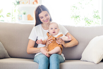 Sticker - Portrait of attractive kind cheerful girl holding playing baby sitting on divan spending free time at light home flat house indoors