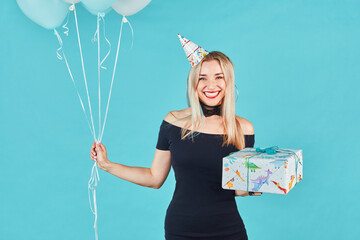 beautiful smiling woman in black dress and birthday hat holding gift box and pastel air balloons aga
