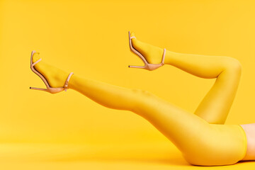 Wall Mural - Long slim legs of young woman in yellow tights and stylish high heels shoes on yellow background.