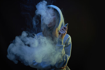 Wall Mural - Portrait of beautiful blonde girl in gray hoodie smokes vape isolated on black studio background, cloud of steam smoke, mini hookah