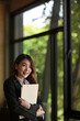 © saltdium - Photo of a young businesswoman standing and holding a document and clipboard over a comfortable meeting room as a background.