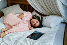 Joyful Female In Pink Sleepwear Lysing At Comfortable Bed With Digital Tablet Technology And Dreaming During Daytime, Cheerful Asian Woman With Closed Eyes Smiling During Sunday Lazy Day In Bedroom