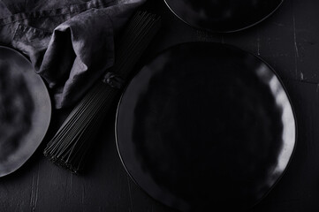 Dark food background with empty black stone board, ceramic plate, raw nero di seppia spaghetti and textile napkin. Free space for text or product. Top view, flat lay. Copy space