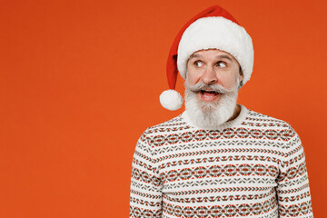 Old shocked surprised stupefied fascinating charming jubilant bearded Santa Claus man 50s wear Christmas hat sweater posing looking aside keeping mouth open isolated on plain orange background studio