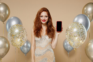 Wall Mural - Young redhead woman in evening dress near balloons hold mobile cell phone with blank screen workspace area isolated on plain pastel beige background studio portrait Celebrating birthday party concept