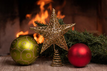 Golden Christmas Star Next To Green And Red Bulbs, Next To The Branch Of An Artificial Tree On A Wooden Table In The Background A Campfire, Christmas Decorative Objects In Studio, Party Celebration