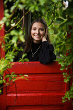 Happy Teenage Girl Leans Through Window Of Bright Red Door Found In The Middle Of An Outdoor Garden