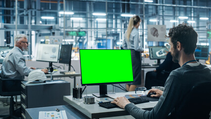 Fototapete - Car Factory Office: Male Automotive Engineer Sitting at His Desk Working on Green Screen Chroma Key Computer. Automated Robot Arm Assembly Line Manufacturing. Medium Shot