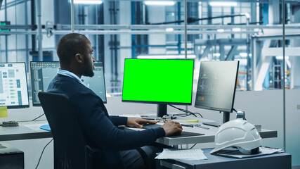 Poster - Car Factory Office: Black Engineer Works on a Computer, Two Monitor Screens Show Chroma Key Green Screen and Monitoring of Automated Robot Arm Assembly Line Manufacturing High-Tech Electric Vehicles