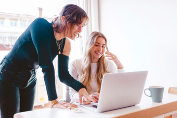 Two young beautiful cheerful women working together with laptop at the office. Entrepreneurs cooperating and discussing about the best business plan in their job at workplace. Using technology.