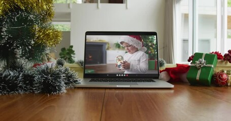 Wall Mural - Caucasian boy with santa hat playing with snow globe on christmas video call on laptop