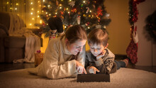 Portrait Of Boy With Mother Warm Wool Sweater Lying On Floor And Using Digital Tablet Computer On Christmas Eve At House