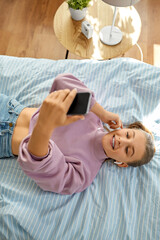 Wall Mural - leisure, children and technology concept - happy smiling girl in earphones with smartphone lying on bed at home