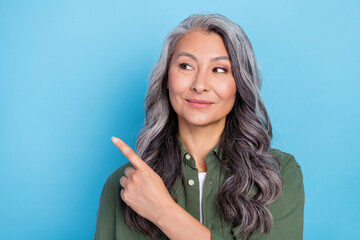 Photo of thoughtful sweet retired woman wear green shirt pointing finger empty space smiling isolated blue color background