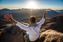 A Man Sits Facing The Rising Sun With Raised Hands On Mount Sinai In Egypt.