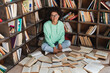 pleased african american student in eyeglasses sitting with crossed legs surrounded by books in library