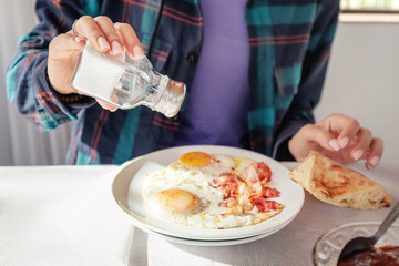 Sprinkles eggs and bacon with salt from a shaker. An excess of sodium chloride mineral can lead to the development of heart and metabolism diseases