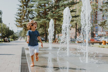 Little Boy Is Running In The Street And Playing With The Water Jets Of A Fountain Spouting From The Ground. Child In Blue T-shirt And White Shorts Is Fooling Around Outdoors. Lifestyle. Space For Text