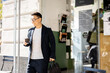 Asian businessman with coffee going from entrance of building. Smiling modern adult successful man wearing suit and glasses with briefcase. City at sunny day