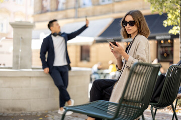 Wall Mural - Young woman and asian man using smartphones on city area with fountain. Concept of city life. Beautiful girl wearing glasses sitting on shair. Guy with headphones. Warm sunny day