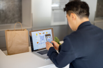 Man using smartphone for payment in online store on laptop on kitchen at home. Concept of online shopping and e-commerce. Idea of healthy and vegetarian eating. Person sit at table with shopping bags
