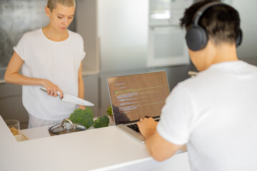 Caucasian woman cooking dish from fresh vegetables while her husband in earphones typing something on laptop. Concept of healthy and dieting eating. Domestic lifestyle.