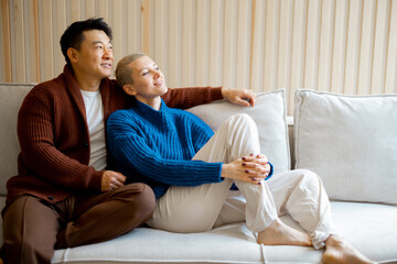 Asian man and young caucasian woman sitting and resting on sofa at home. Concept of relationship and spend time together. Idea of domestic lifestyle. Smiling and pensive multiracial couple look away